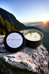 An open tin of The Lather and The Wick's "A New Day - Wax Melt," crafted from sustainable soy wax, sits on a rocky surface with mountains and a valley in the background. The sunrise illuminates the sky, casting light on the fresh and clean melts and their reusable metal tin amidst the natural scenery.