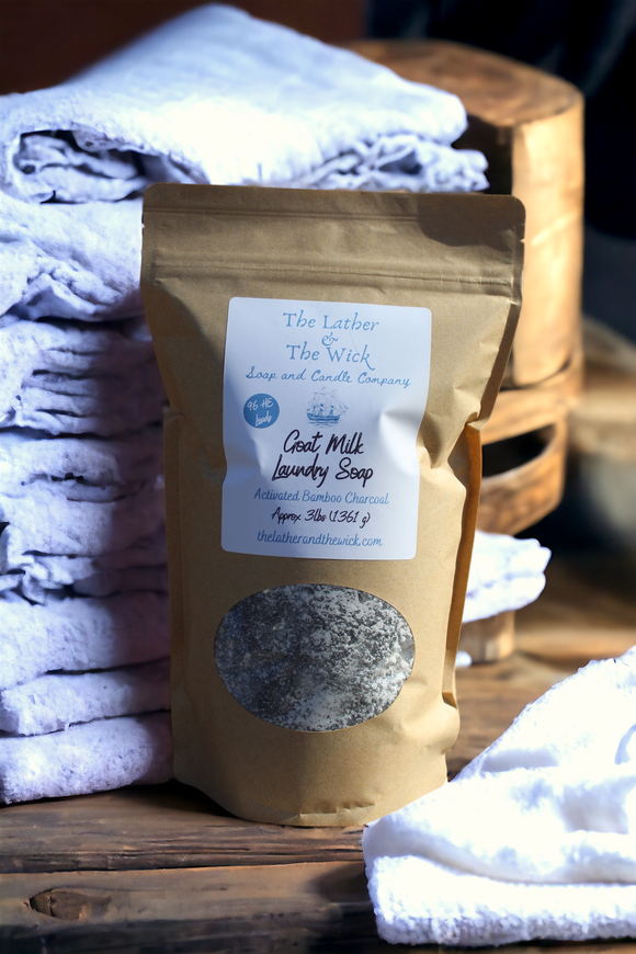 A brown kraft paper bag of 100% Natural Goat Milk Laundry Soap - 4 Scents from The Lather and The Wick sits on a wooden surface. This chemical-free detergent, containing activated bamboo charcoal, weighs 12.9 oz (365g). Surrounding the bag are folded white towels and wooden accents.