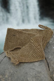 An All Natural Back Scrubber from The Lather and The Wick is casually draped over a large rock in the foreground. In the background, a waterfall cascades down, creating a serene and natural scene. The focus is on the texture of the biodegradable back scrubber against the smooth rock with the blurred waterfall behind it.