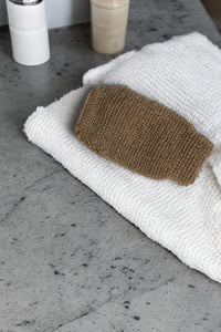 A white, textured towel is folded on a gray speckled countertop. On top of the towel is an All Natural Bath Mitt by The Lather and The Wick, perfect for exfoliating and cleaning. In the background, there are two small containers, one white and one beige.