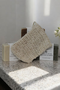 Three premium goat milk soap bars labeled "Lavender" and "Water Nymph" are displayed on a speckled countertop. The All Natural Soap Saver by The Lather and The Wick is placed on top of the soaps, while a small bunch of white flowers adds a delicate touch in the background.