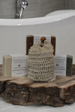Four bars of soap with various natural ingredients, labeled as "Lavender Fields," "River Nymph," and "Detox Goat Milk Soap," from The Lather & The Lick Soap & Candle Co. sit on a rustic wooden platter in front of a white bathtub. A Pre-filled All Natural Soap Saver from The Lather and The Wick is positioned in front.