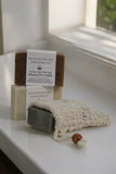 A set of three soap bars in neutral shades, with one bar partially inside The Lather and The Wick's All Natural Soap Saver. The middle soap is labeled "Dragon's Fire, Premium Goat Milk Soap" with ingredients. The soaps are displayed on a white surface near a window.