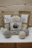 This image displays two packages of Goat Milk Laundry Soap and a sack of The Lather and The Wick 100% Wool Dryer Balls - Set of 6. Three wool dryer balls are placed in front of the sack, with the two soap packages behind it.