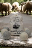 A drawstring bag labeled "The Lather and The Wick, 100% Wool Dryer Balls - Set of 6" is placed on a stone surface. Six wool dryer balls, in shades of gray and cream, are scattered around the bag. In the background, sheep are grazing on a road.